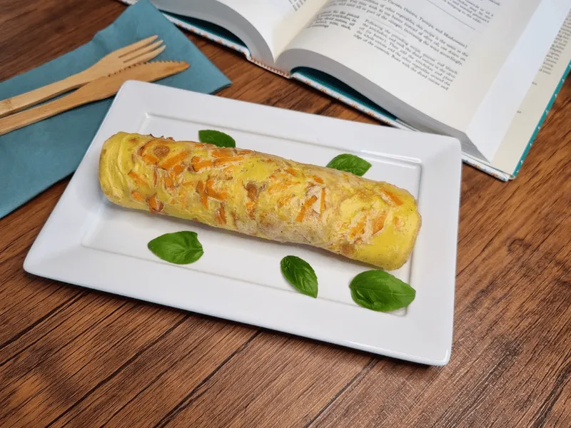 Crepioca enrolada com cenoura ralada aparente na massa, sobre travessa retangular branca decorada com folhas de manjericão, ao lado de talheres de bambu e um livro aberto