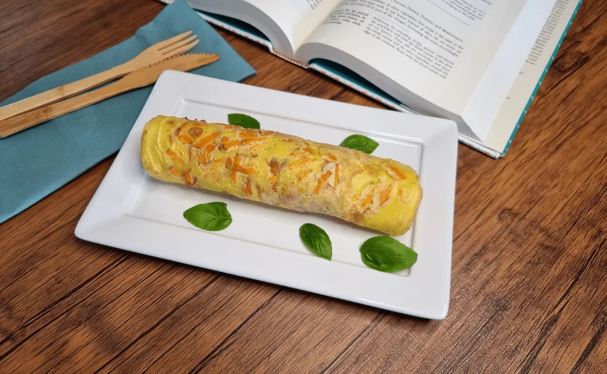 Crepioca enrolada com cenoura ralada aparente na massa, sobre travessa retangular branca decorada com folhas de manjericão, ao lado de talheres de bambu e um livro aberto