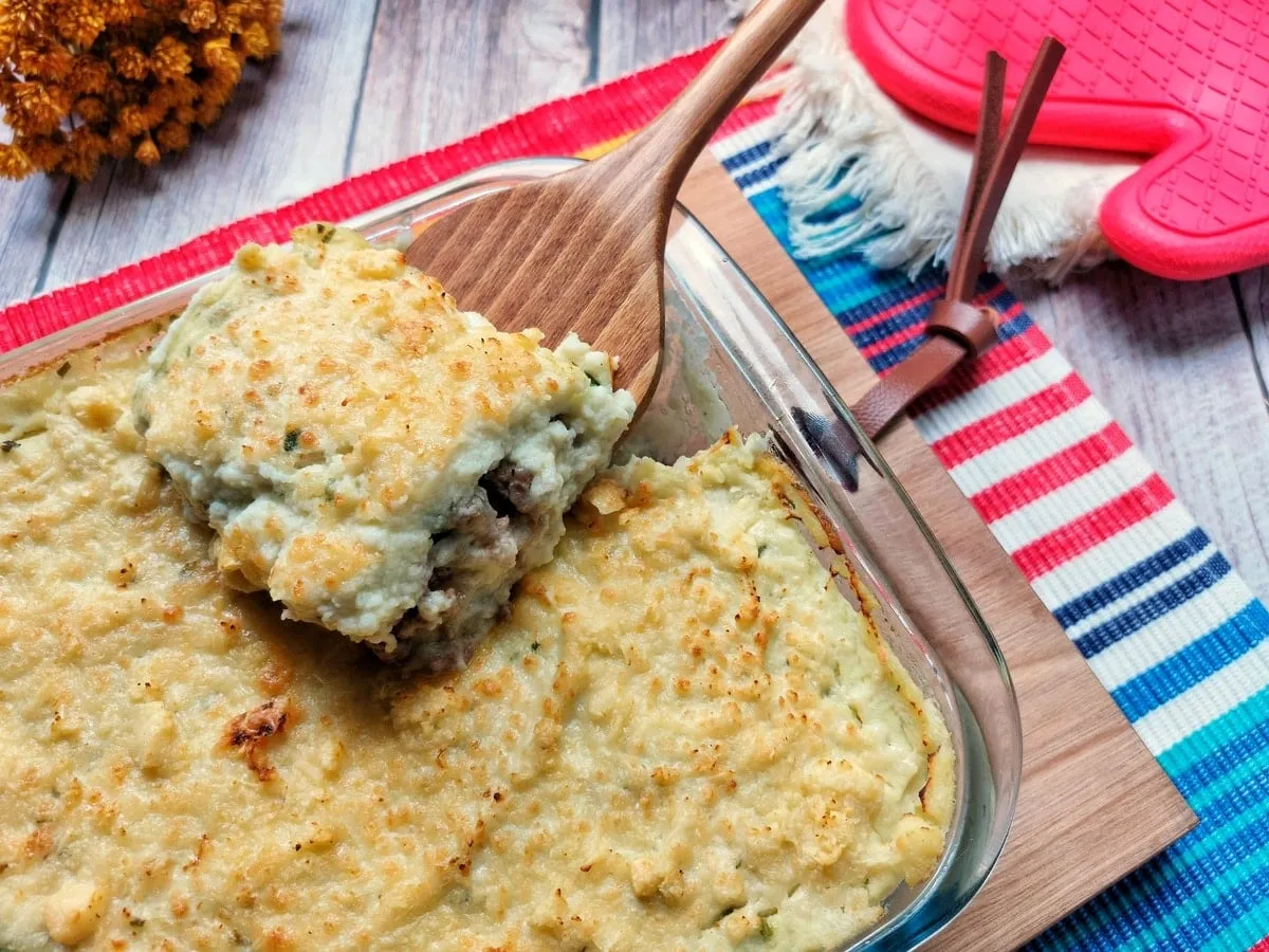 Refratário de vidro com escondidinho gratinado de purê claro, com uma espátula de madeira erguendo uma porção que mostra a carne moída por dentro, sobre jogo americano listrado