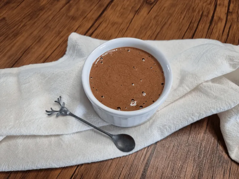 Ramequim branco com mousse de chocolate cremosa sobre um pano de algodão claro, ao lado de uma colherzinha de metal, em mesa de madeira
