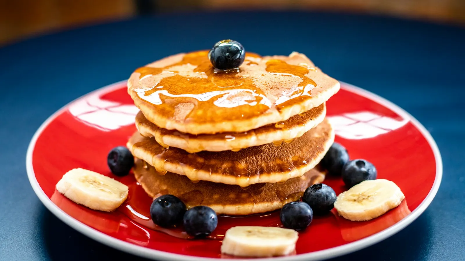 Pilha de panquecas com mel, mirtilos e rodelas de banana em prato vermelho