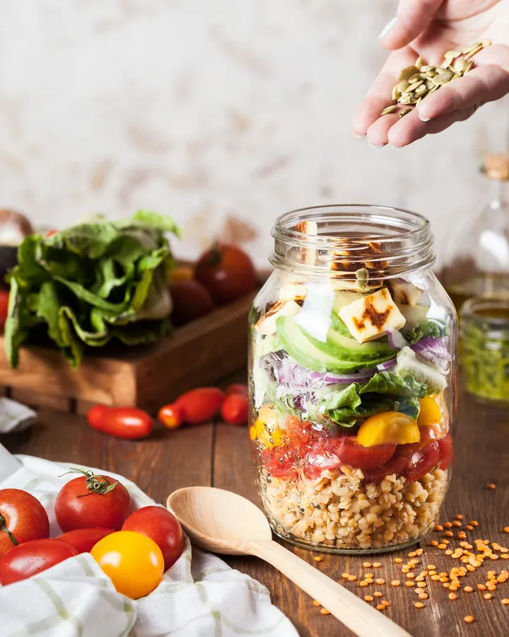 Salada montada em pote de vidro sobre mesa de madeira, com tomates e folhas ao redor