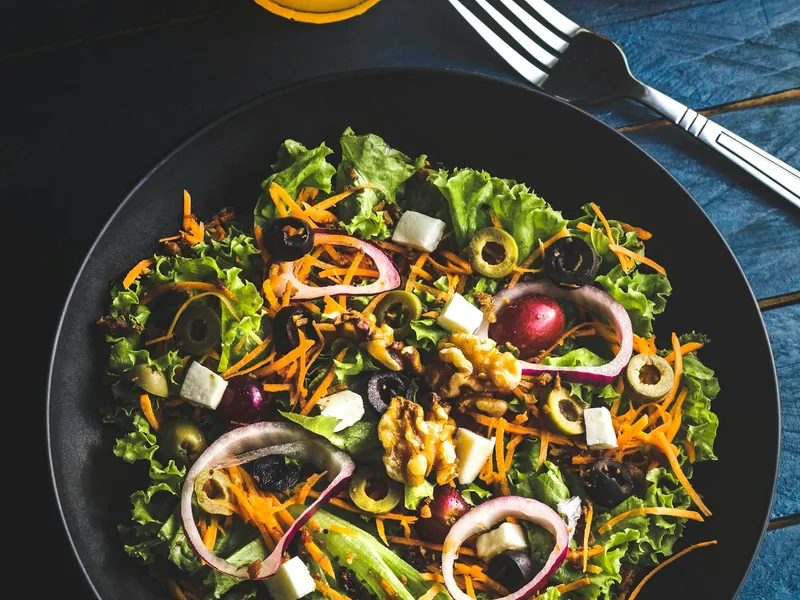 Salada de alface, cenoura ralada, nozes, azeitonas e queijo branco em prato escuro, com copo de suco de laranja